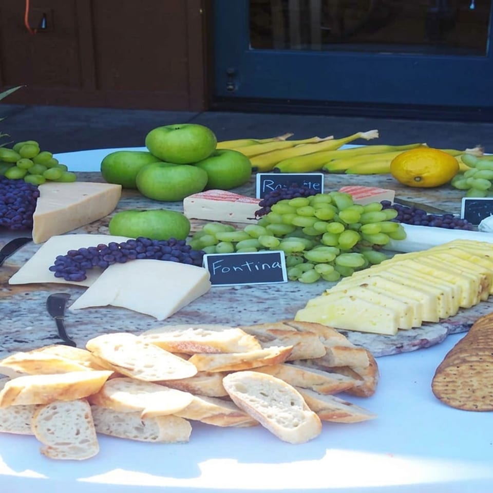 Outdoor cheese and fruit table featuring sliced cheeses, grapes, berries, pineapple, apples, crackers, and assorted breads.