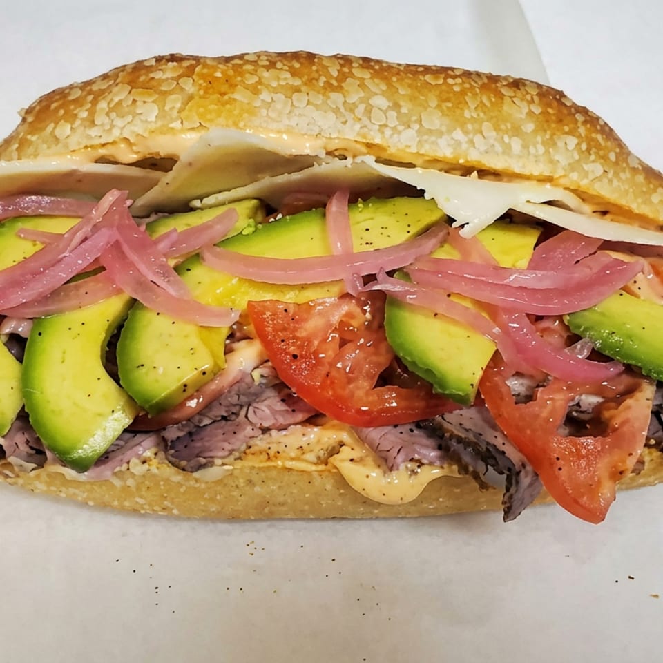 Large seeded sandwich filled with sliced roast beef, avocado, tomato, pickled onions, melted cheese, and creamy sauce.